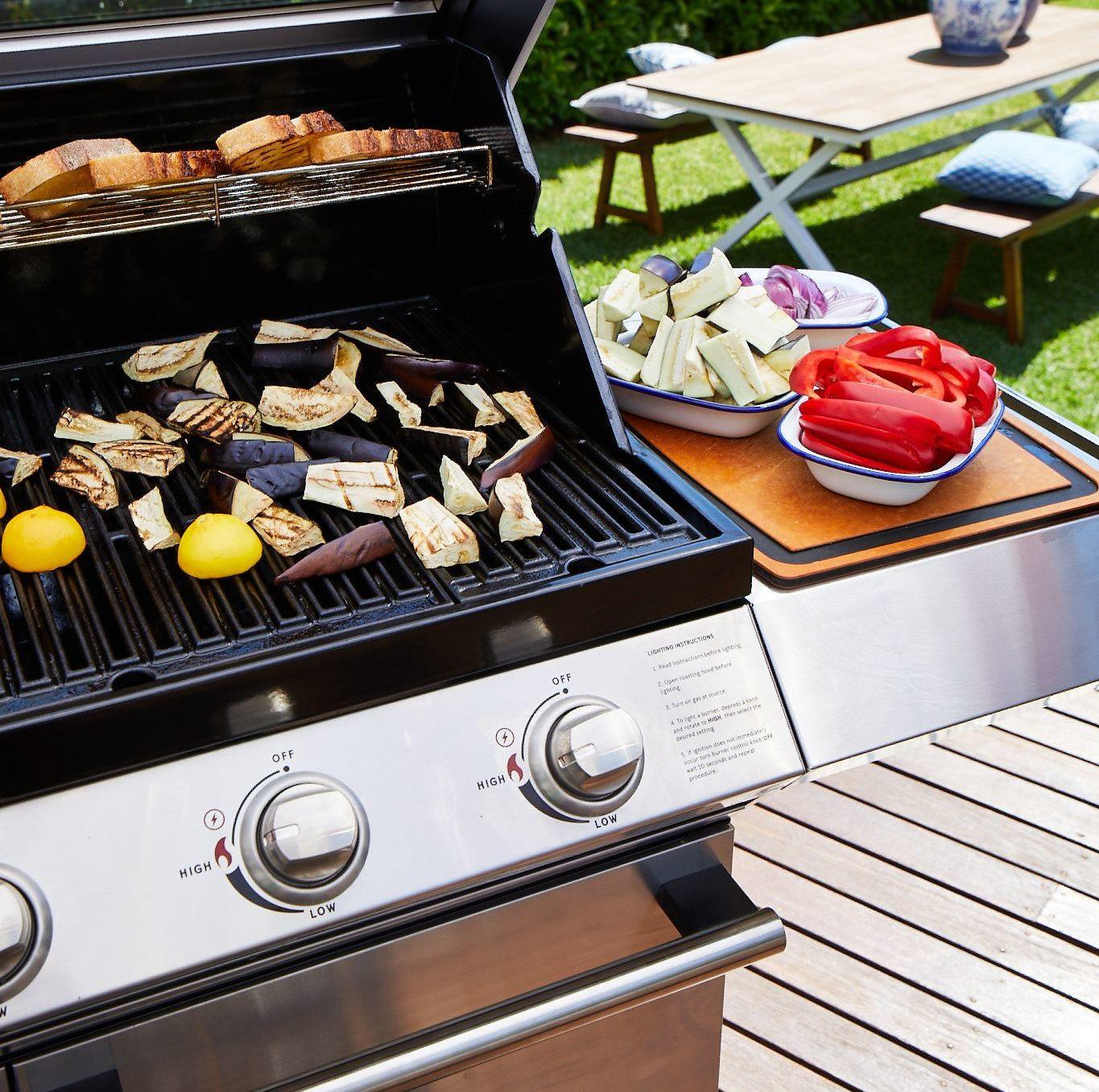 A Brisks BeefEater 1200E Series 3 Burner BBQ & Trolley on a wooden deck with sliced vegetables like eggplants, red peppers, and lemons cooking on the grates sets the perfect scene for outdoor living. Additional bowls of cut vegetables are placed on a side table next to the BBQ. A picnic table with white bench seating is in the background.