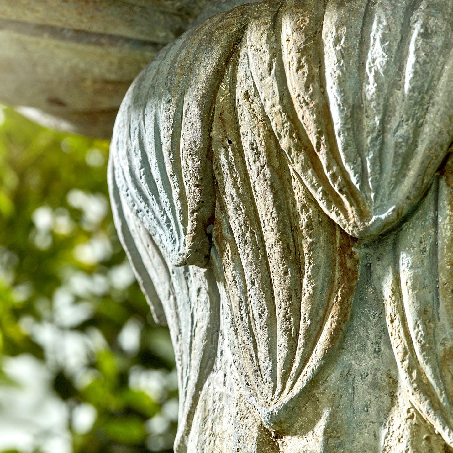A detailed view of the textured stone sculpture from La Hacienda's Lioness Fountain showcases intricate grooves that mimic flowing drapery. The piece is beautifully set against a softly blurred green foliage background, creating an outdoor ambiance.