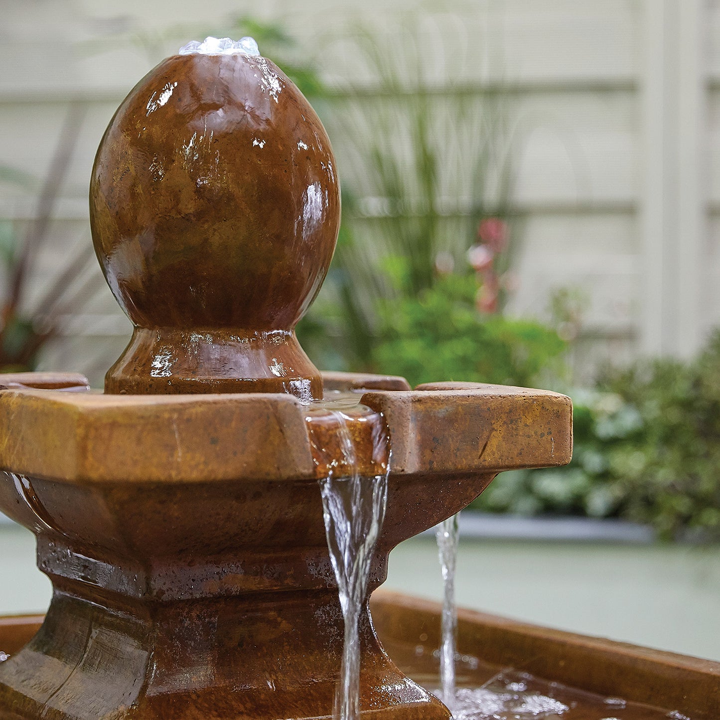 A close-up of the La Hacienda's Odyssey inc LEDs, a decorative glass fibre reinforced concrete fountain, shows water gently flowing over its rounded top. Enhanced by subtle LED lights, the background features blurred greenery and a light-colored wall, creating a serene garden setting.