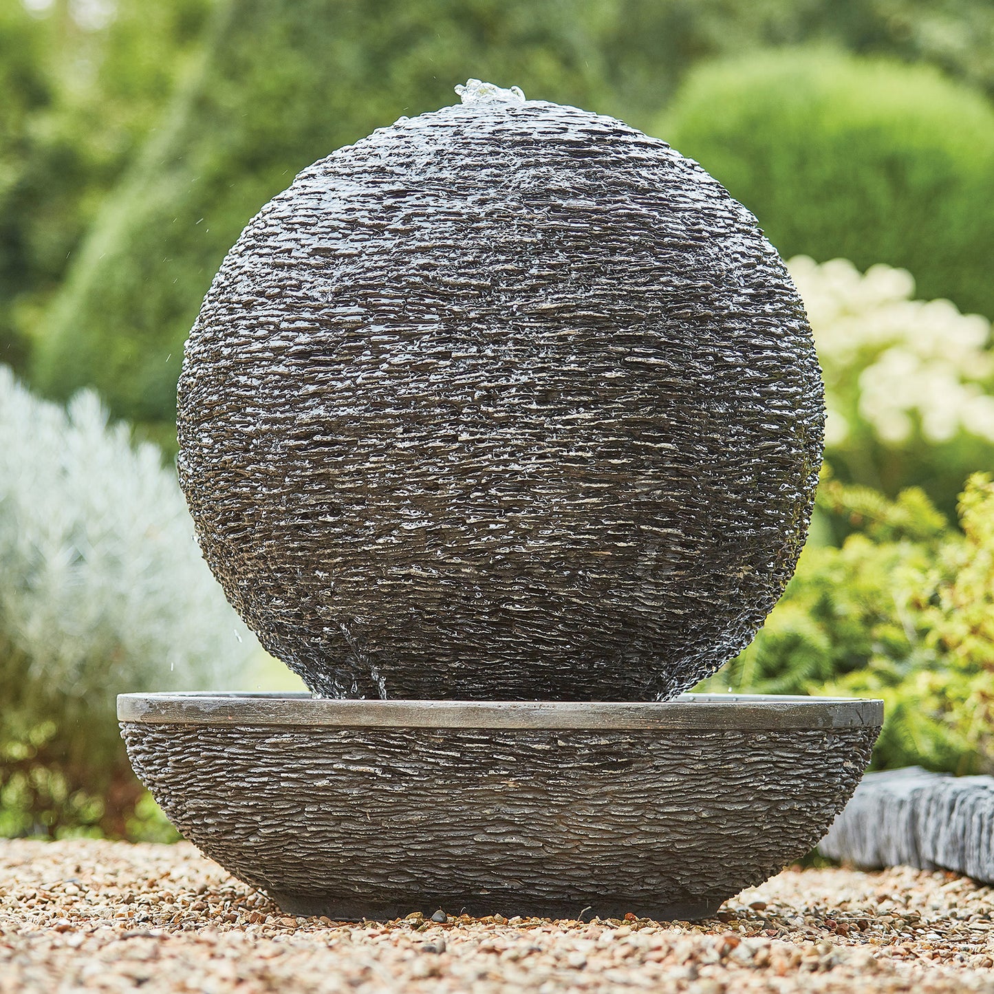 The Mysterious Moon by La Hacienda, a large spherical stone water feature made from glassfibre reinforced concrete, is positioned on a matching basin surrounded by gravel and lush greenery. This self-contained piece allows water to cascade down its textured surface, enhancing the soothing outdoor aesthetic.