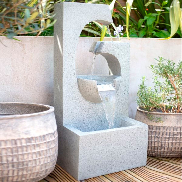 The La Hacienda Ango Falls is a contemporary outdoor waterfall with a sleek geometric design. Water cascades from a circular cutout into a rectangular basin, framed by potted plants, to create a tranquil garden atmosphere. It serves as an impressive modern statement for any garden.