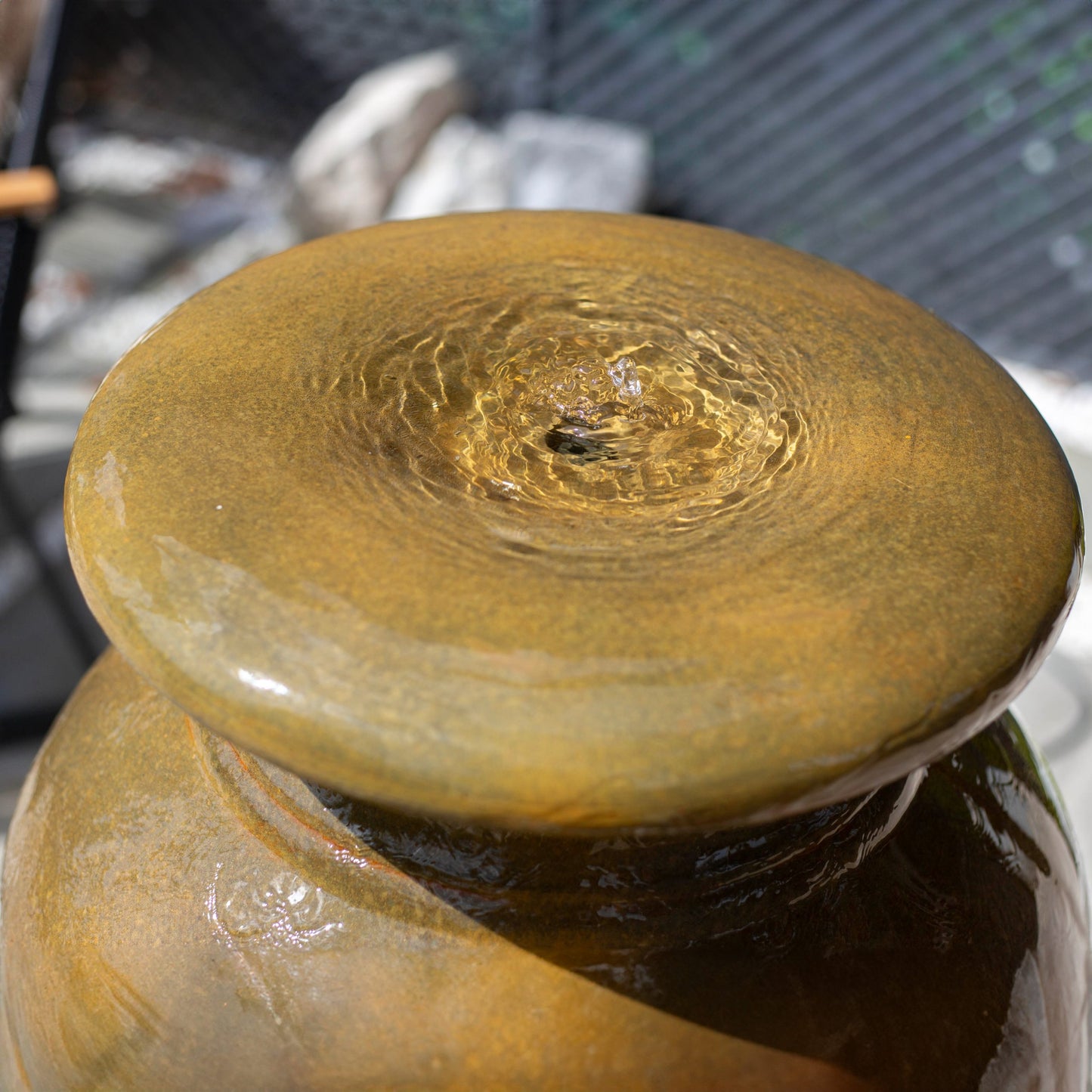 Close-up of the La Hacienda Athenian Vase water fountain, with a smooth, brownish-gold glaze. Water gently bubbles from a small opening at the top, creating ripples that glisten in the sunlight. A blurred background includes rocks and a fence.