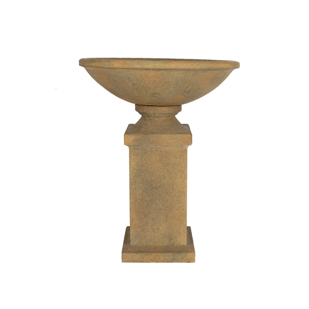 A stone birdbath featuring a wide, shallow bowl gracefully rests on La Hacienda's Roman Pedestal. This garden feature exudes a rustic, weathered look that reflects its natural stone composition. The design is simple yet elegant, imparting timeless charm to any outdoor setting.