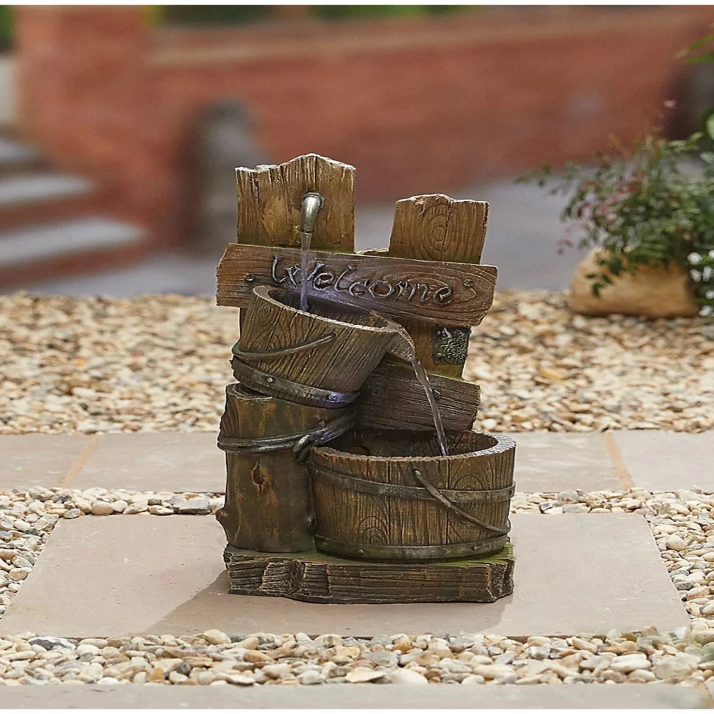 A rustic wooden garden fountain, the Fence Post Pours inc LEDS by La Hacienda, is a delightful addition to your garden decor ensemble. It features a "Welcome" sign with water gently cascading into a barrel. Positioned on a pebble and stone surface, it beautifully enhances the blurred greenery and stairs in the background.