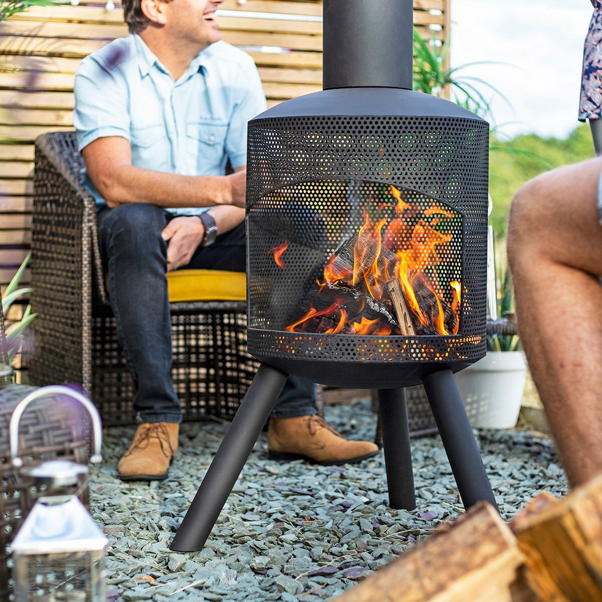 Two people sit around a lively fire in the La Hacienda Santana Fireplace, with its contemporary design making a statement. The person on the left wears a light blue shirt and jeans, while the other is dressed in shorts. They're seated on a gravel patio with a wooden screen and plants as the backdrop.