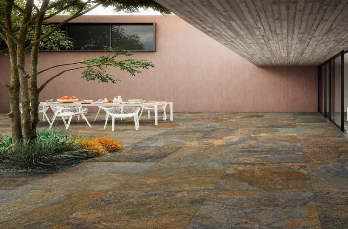 A contemporary outdoor patio features Brisks' slip-resistant Copper Moon Porcelain Paving tiles, a dining table with white chairs, and a small garden area with a tree and plants on one side. The space is partially covered by a concrete overhang, adding sophistication to the outdoor setting.