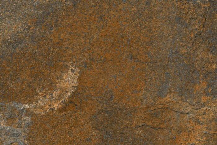 Close-up of a rough, rust-colored textured surface with patches of brown and dark gray. This slip-resistant surface appears uneven and natural, perfect for outdoor areas. Resembling aged stone or weathered metal, it echoes the elegance of Brisks Copper Moon Porcelain Paving tiles.
