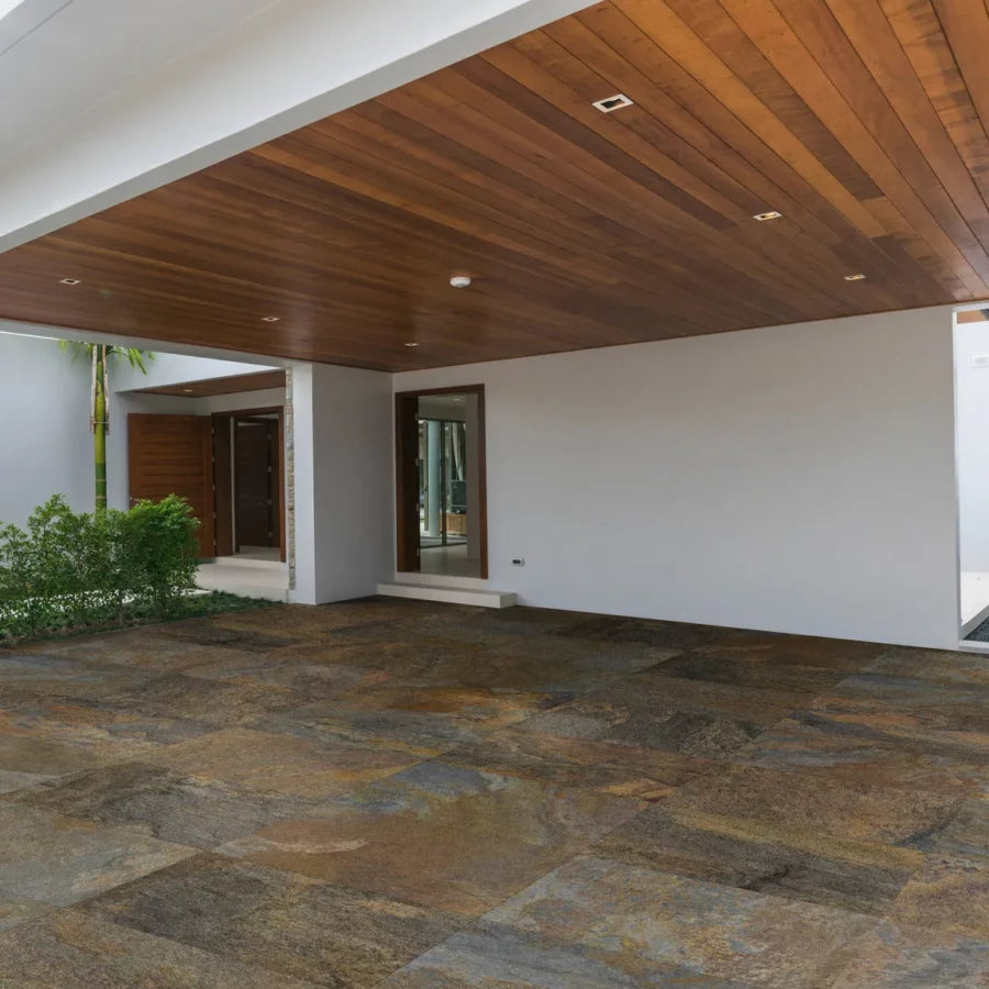 A modern architectural entrance showcases a wooden overhang and Brisks Copper Moon Porcelain Paving for slip-resistant flooring, accompanied by a sleek white wall and reflective glass doors. Lush greenery lines the walkway, enhancing the minimalist design of this chic outdoor area.