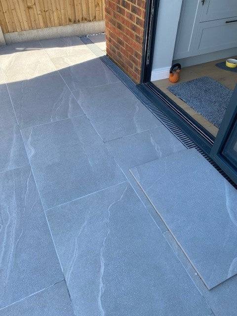 A partially tiled outdoor patio showcases large, light gray Kiara Grey Porcelain Paving from Brisks, with some tiles still unaligned and renowned for their exceptional durability. This slip-resistant surface transitions into an interior space through a partially open sliding glass door. In the background, a wooden fence and brick wall are visible.