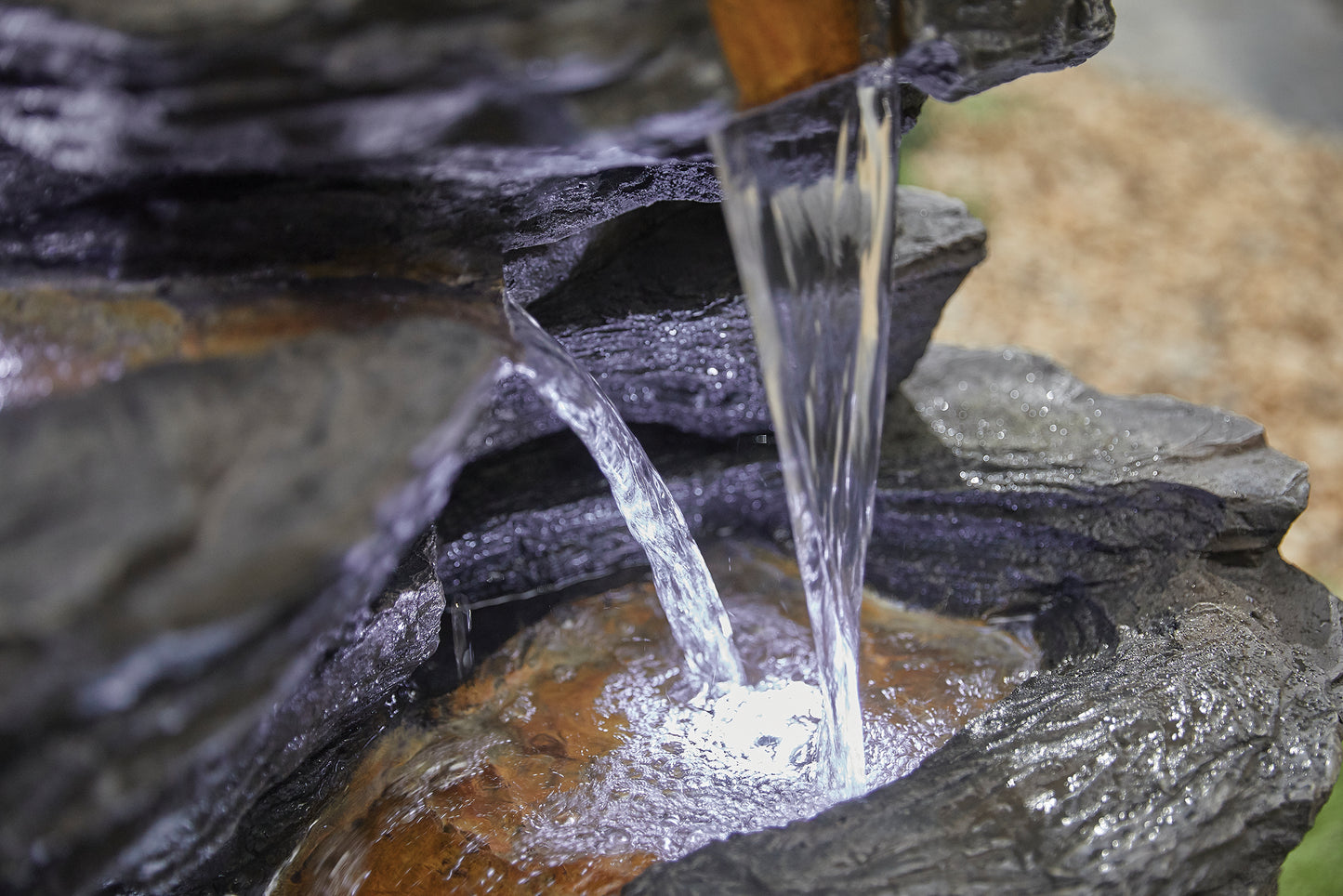 A close-up of water cascading over dark, textured rocks into a small pool evokes the serene and natural ambiance of La Hacienda's Hinoki Springs inc LEDs. The gentle flow and smooth surface of the water contrast with the rugged stone, enhancing this tranquil water feature.
