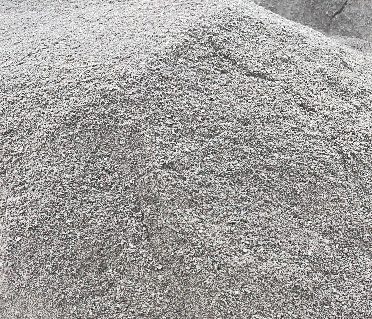 A large mound of fine gray Grano Dust by Brisks is clustered together, forming uneven peaks and slopes. The gravel, commonly used as a sub-base layer, consists of small, coarse particles of varying shades of gray, giving a textured, slightly rough appearance.