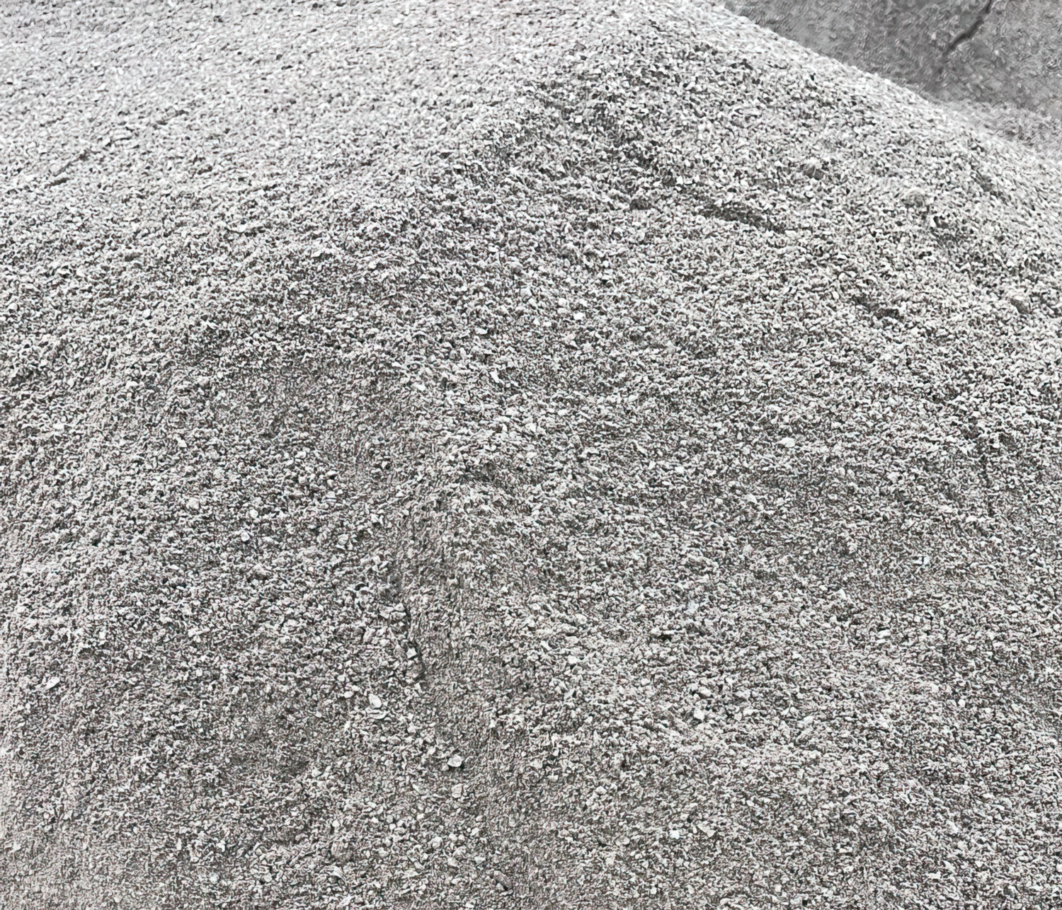 A large mound of fine gray Grano Dust by Brisks is clustered together, forming uneven peaks and slopes. The gravel, commonly used as a sub-base layer, consists of small, coarse particles of varying shades of gray, giving a textured, slightly rough appearance.