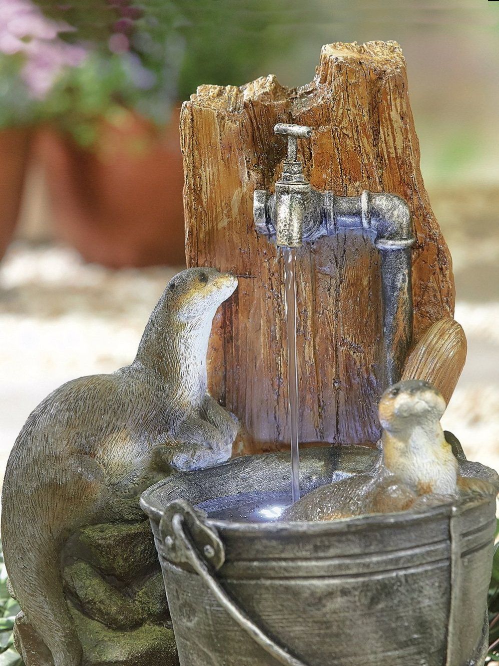The Otter Pools inc LEDS by La Hacienda is a decorative outdoor fountain featuring playful otter sculptures; one gazes up at the water flowing from an old-fashioned faucet into a metal bucket, while the other appears to be sitting in it. Enhanced with LED lights, this self-contained setup is charmingly placed on a rocky surface.