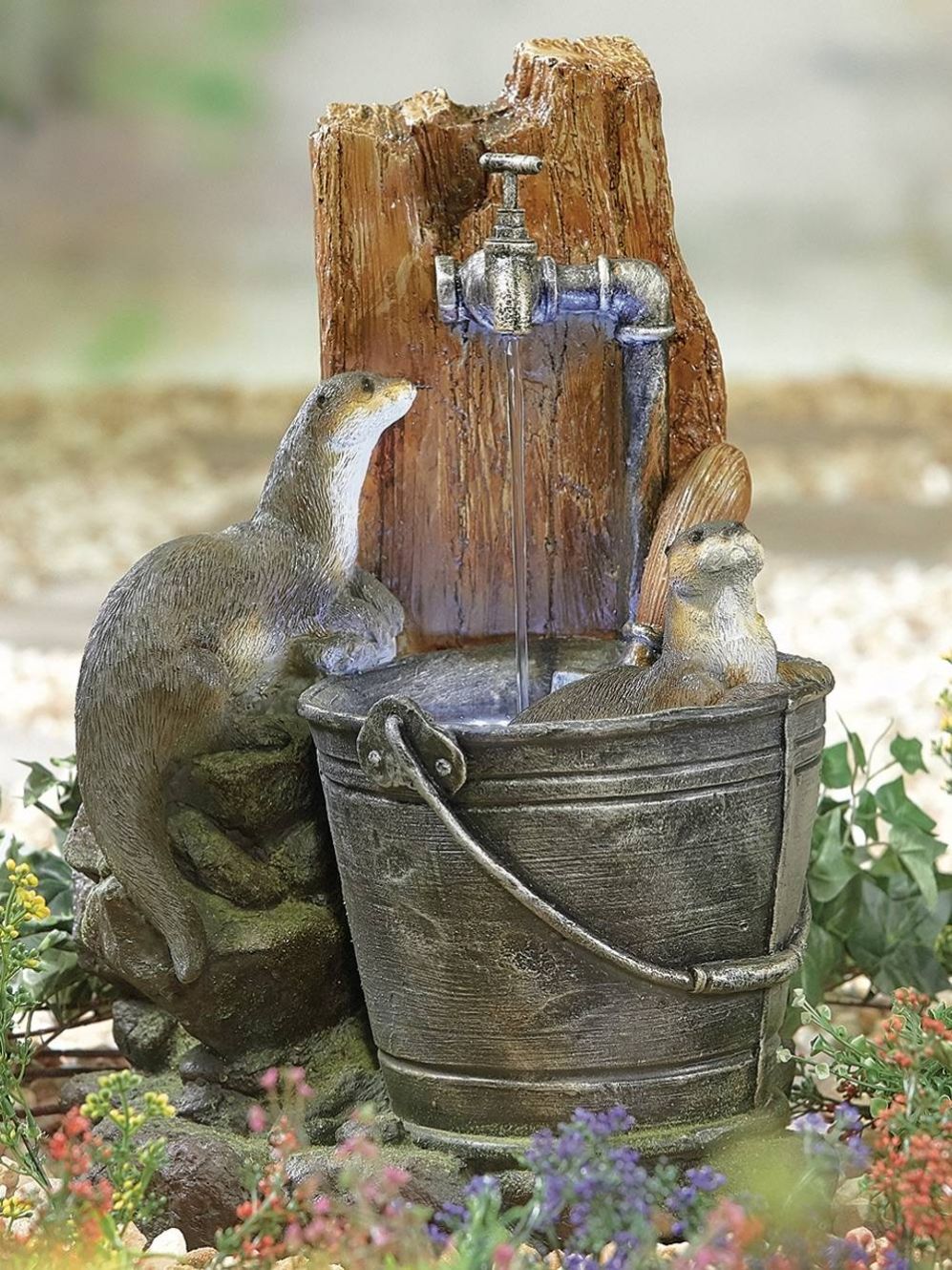 The "Otter Pools inc LEDS" by La Hacienda is a whimsical garden sculpture featuring playful otters at a rustic water tap. One otter stands next to a wooden post with a faucet, while the other sits in a metal bucket filled with water. Surrounded by colorful flowers, this self-contained setup enchants both day and night with its charming LED lights.