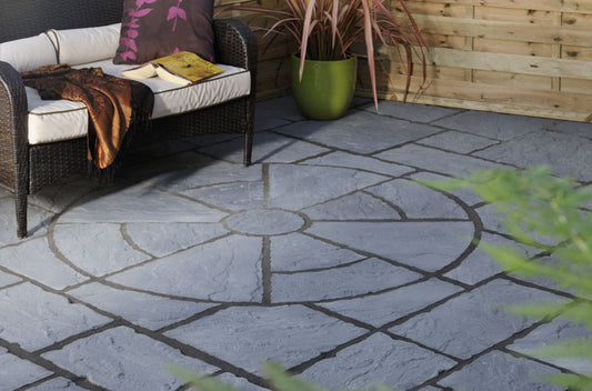 A cozy patio with a wicker sofa adorned with pillows, a throw blanket, and an open book. The floor features the Brisks Welsh Slate Rectory Circle Patio Kit by Bowland Stone, surrounded by rectangular stones. A green potted plant adds a touch of nature. Wooden panels line the perimeter, enhancing the garden landscaping charm.