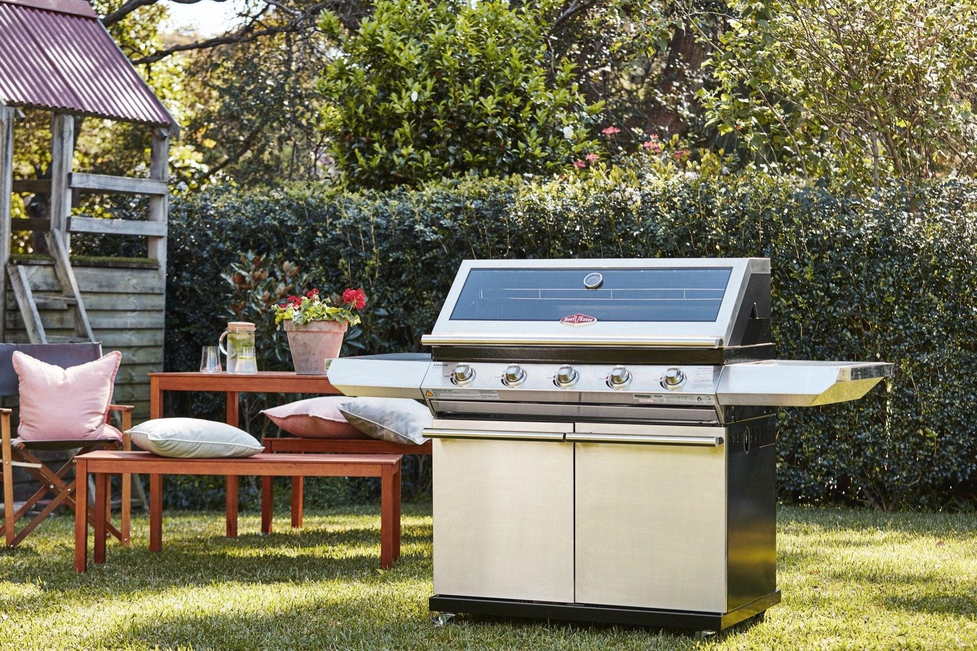 Outdoor scene with a Brisks BeefEater 1200S Series 4 Burner BBQ & Trolley featuring multiple knobs and a closed lid, perfectly suited for BBQs, placed on a grassy lawn. To the left, there's a wooden picnic bench with a potted plant, pink and white cushions, and a wooden playhouse near green hedges under the clear sky—ideal for modern outdoor living.
