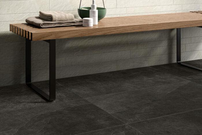 A modern bathroom features a wooden bench with black metal legs against a gray tile wall. On the bench are folded towels and bottles, with a green bowl beside them. The floor has large, dark gray Frame Groove Porcelain Paving Slabs from Brisks, adding both style and durability to the space.