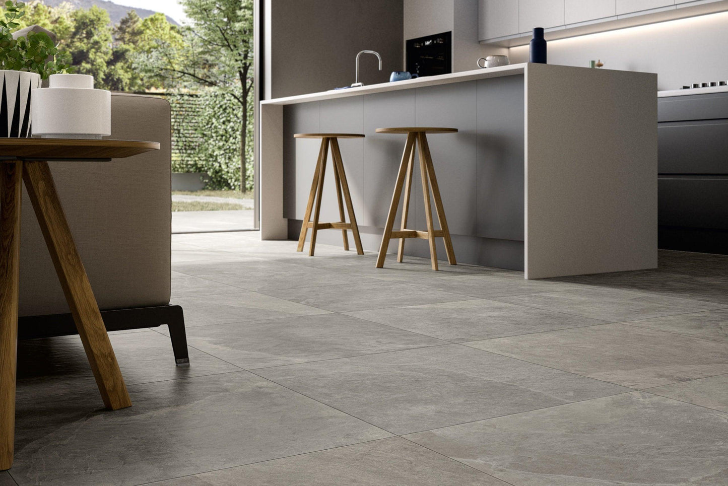 A modern kitchen with a large island featuring two wooden barstools. The floor is paved with Brisks Frame River Porcelain Paving Slabs, offering optimal coverage. The space is bright with natural light, and there is a view of a green yard through glass doors in the background.