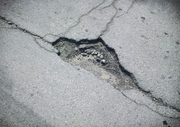 An asphalt road with a large pothole in the center awaits Brisks Cold-Lay Patching Tarmac. The edges of the pothole are cracked, revealing exposed gravel and dirt. The surrounding road surface appears worn and weathered, calling for immediate temporary road repair.