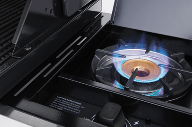 Close-up of a lit gas burner on a sleek, modern stovetop. The blue flame dances around the burner, encircled by a black grate. This stylish setup effortlessly complements any outdoor kitchen featuring premium appliances like the Brisks BeefEater 1200E Series 4 Burner BBQ & Trolley for ultimate culinary experiences.