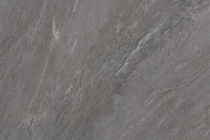 A close-up of a slab of gray stone with natural texture and veining. The Charcoal Grey colour surface displays various shades of gray with patches of lighter and darker streaks running across it, creating an abstract and rugged appearance, ideal for outdoor durability in Brisks Ultra Aspen Anthracite Porcelain Paving Tiles.
