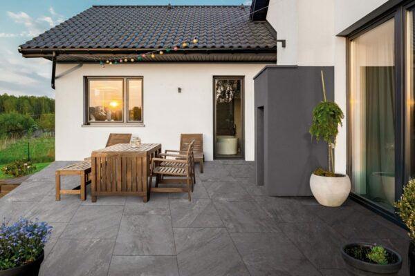 A modern outdoor patio with Brisks Ultra Aspen Anthracite Porcelain Paving Tiles, a wooden table and chairs, and potted plants. The white house features a large window with a sunset reflection, and hanging string lights add a cozy atmosphere. A green lawn and trees are visible in the background.