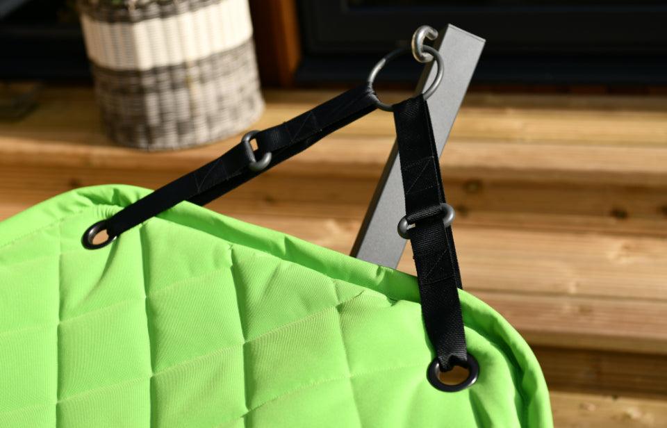 A durable and comfortable green padded fabric is attached to black straps with metal rings. The straps are connected to an outdoor wooden structure, likely part of a stair or deck. A striped basket is partially visible in the background, indicating an easy setup for the Brisks Extreme Lounging B-Hammock.