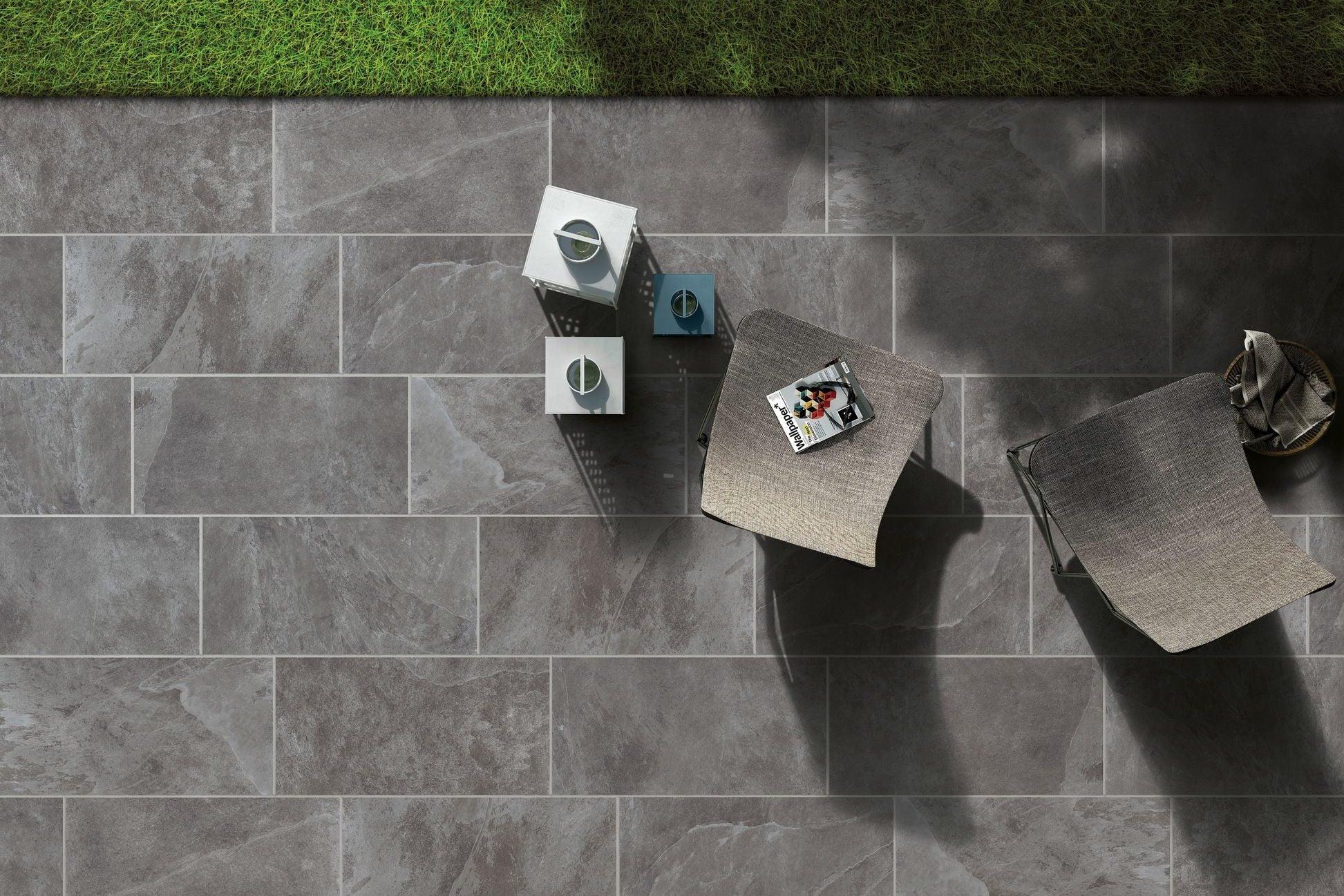 A top-down view of a gray tiled patio featuring two chairs, a side table with a small tray holding a cup, and two decorative lanterns. The top of the image shows a strip of green grass next to the Brisks Frame Groove Porcelain Paving Slabs. Shadows are cast over parts of the patio, enhancing its textured surface.