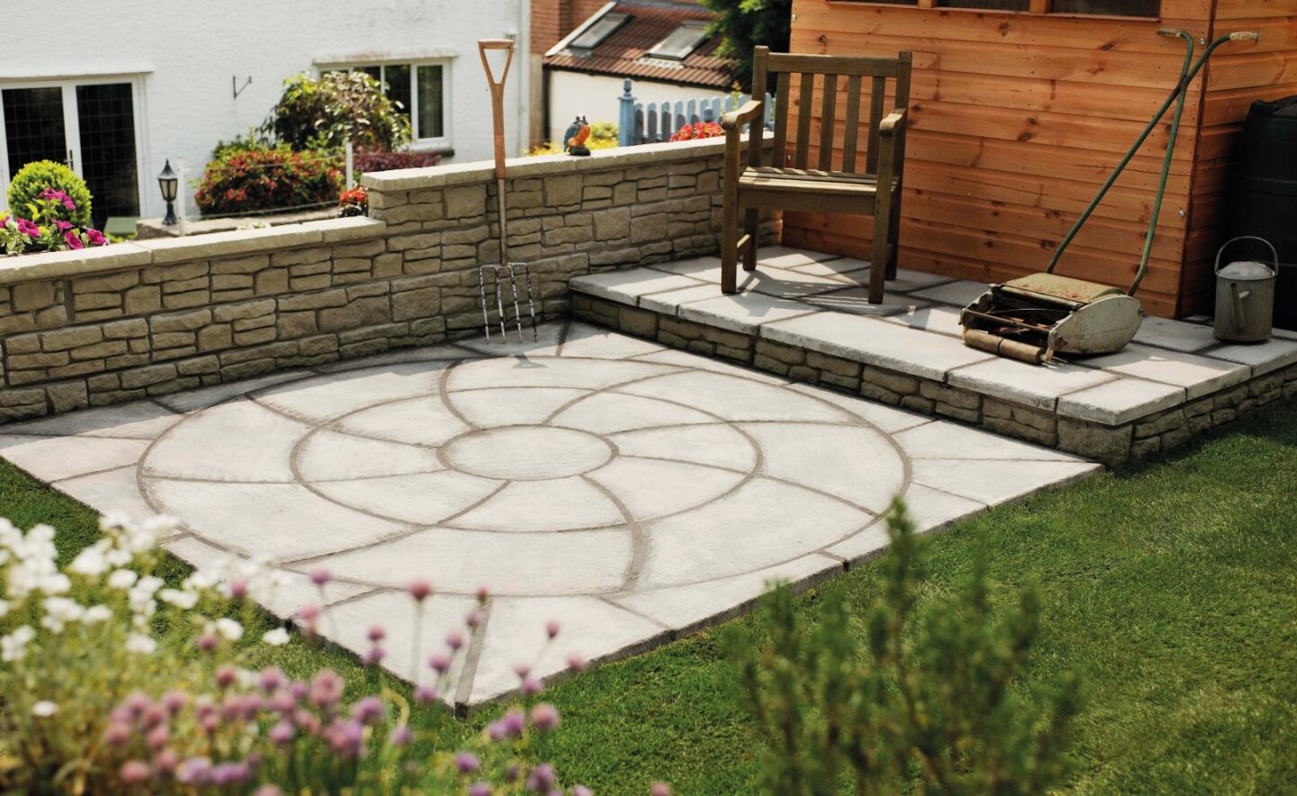 A well-maintained backyard with a circular Weathered Slate Catherine Wheel Kit patio by Brisks in the foreground, bordered by neatly trimmed grass. A wooden bench, spade, and garden tools are on a raised stone platform near a small wooden shed. Flower pots and a stone wall are in the background.