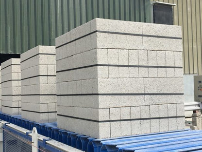 Brisks' 100mm Solid Dense 7.3N Blocks, neatly strapped in uniform square stacks, are set on blue pallets against a corrugated metal wall—ideal for construction projects due to their reliable strength.