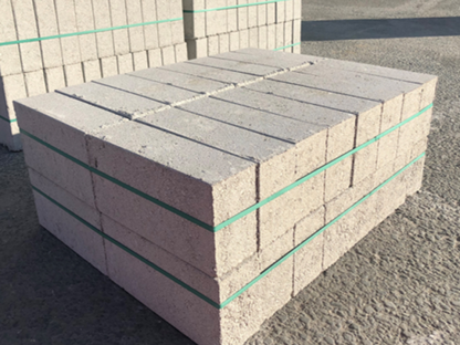 A large stack of Brisks 140mm Solid Dense 7.3N Blocks, known for their reliable strength in construction, is neatly piled in a square shape and secured with green plastic straps. The stack is on a paved surface with more stacks visible in the background.