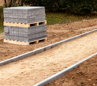 A stack of Brisks 20mm All In Ballast Aggregate, ideal for various construction applications, sits on wooden pallets beside a partially constructed walkway. The walkway has a base layer of sand and is outlined by gray concrete curbs. The surrounding area shows bare earth and some greenery.
