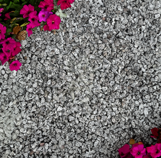 Small Brisks 20mm Black and White Granite Chippings cover the ground's surface with vibrant pink flowers bordering the top left and bottom right corners. The flowers add a splash of color to the otherwise monochromatic rocky background.