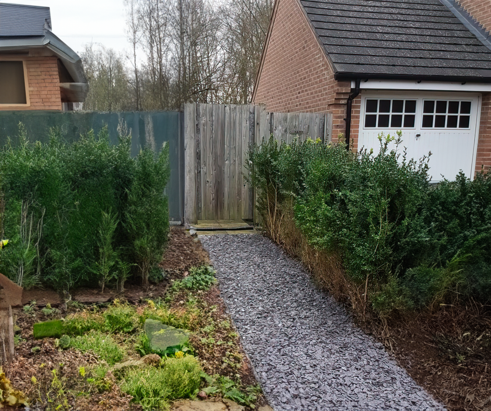 A garden features a gravel pathway bordered by small green bushes and various plants. At the end of the path, edged with 20mm Mixed Blue & Plum Slate Chippings from Brisks, a wooden gate leads to a brick garage with a gray roof and a white door. In the background, trees enhance this landscaping haven.