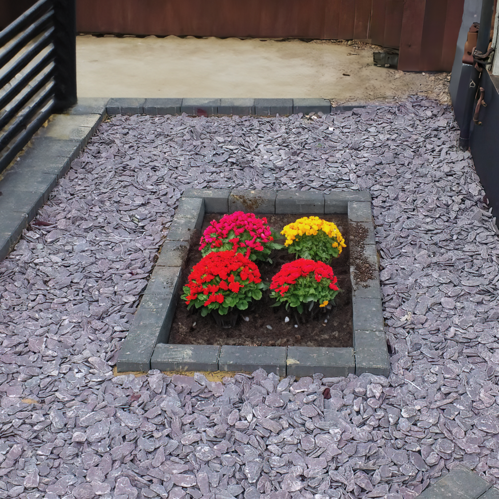 A small garden area featuring a rectangular flower bed with vibrant red and yellow flowers. The bed is bordered by dark stone bricks and surrounded by Brisks 20mm Mixed Blue & Plum Slate Chippings. The background includes a wooden fence and a paved area, adding to the overall landscaping charm.