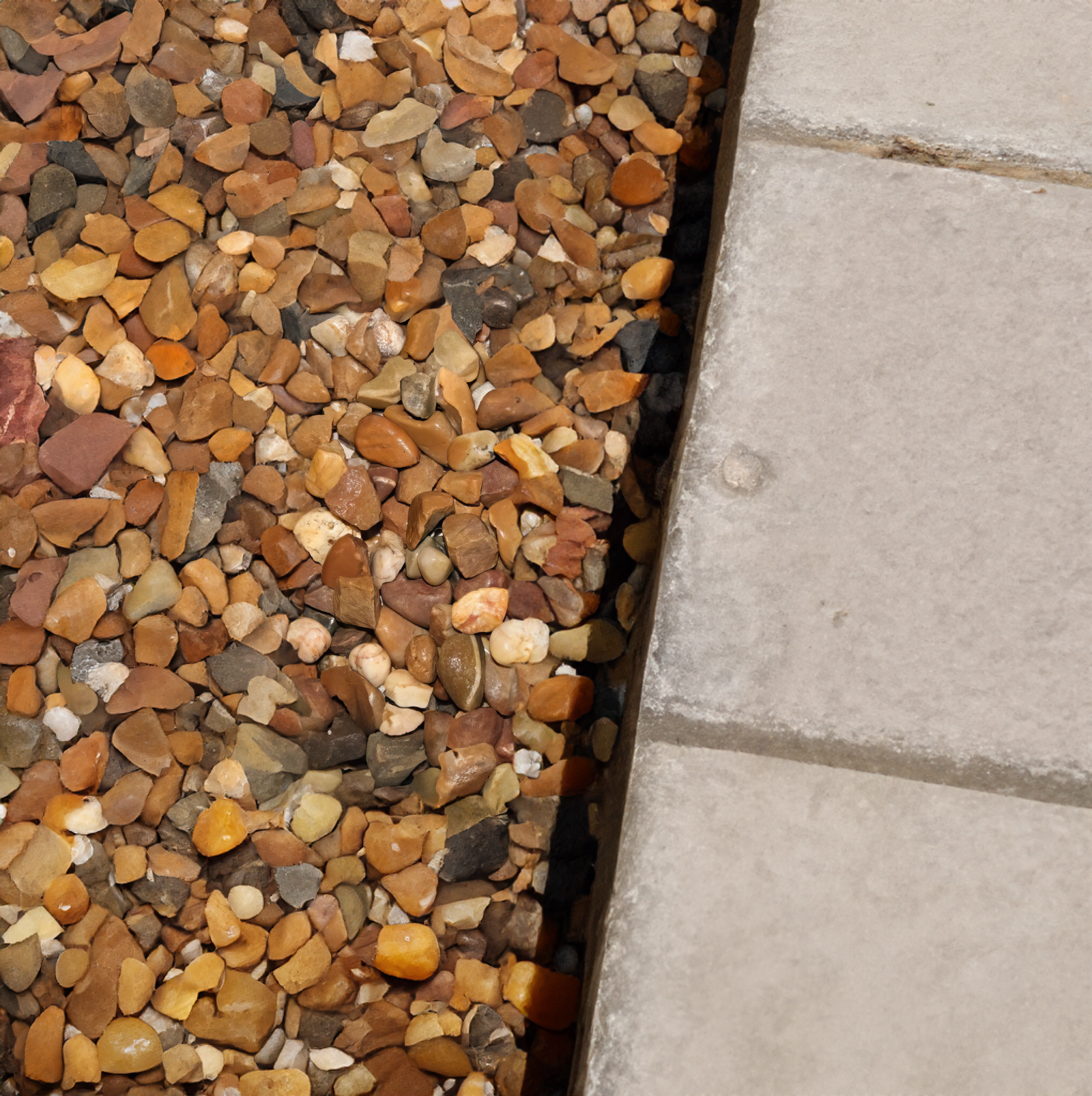 A close-up of small, multicolored pebbles, possibly 20mm Trent Valley Gravel, next to a concrete path. The Brisks 20mm Trent Valley Gravel in brown, tan, and orange hues contrasts with the smooth gray tiles, adding charm to the outdoor landscape.