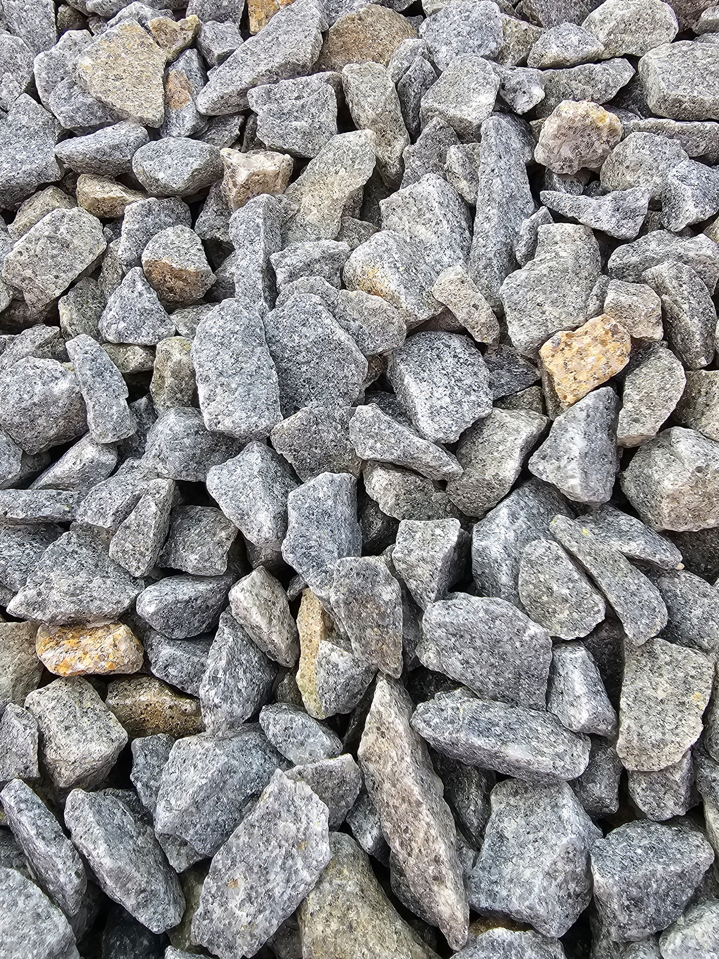 A close-up image showcases a pile of Brisks' 20mm Cornish Granite driveway chippings, featuring uneven, jagged edges. The stones exhibit slight variations in shade and size, offering a rough texture ideal for landscaping, with some displaying patches of brownish discoloration.