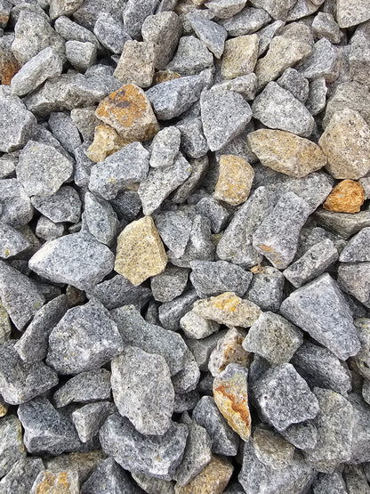 A detailed view of 20mm Cornish Granite from Brisks displays a collection of irregularly shaped gray stones interspersed with lighter, yellowish pieces. Ideal for landscaping or use as driveway chippings, these rocks offer a varied range of sizes and textures, resulting in an appealingly diverse surface.