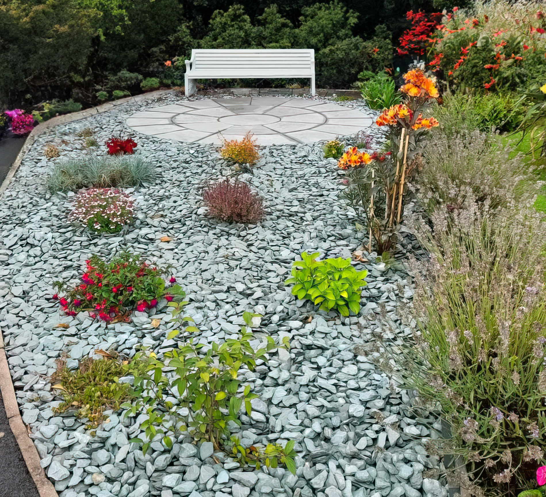 A garden with vibrant flowers and plants is bordered by light gray pebbles and Brisks' 40mm Green Slate Chippings. A white bench rests on a circular stone area, framed by lush greenery and red flowers, highlighting exquisite landscape design.