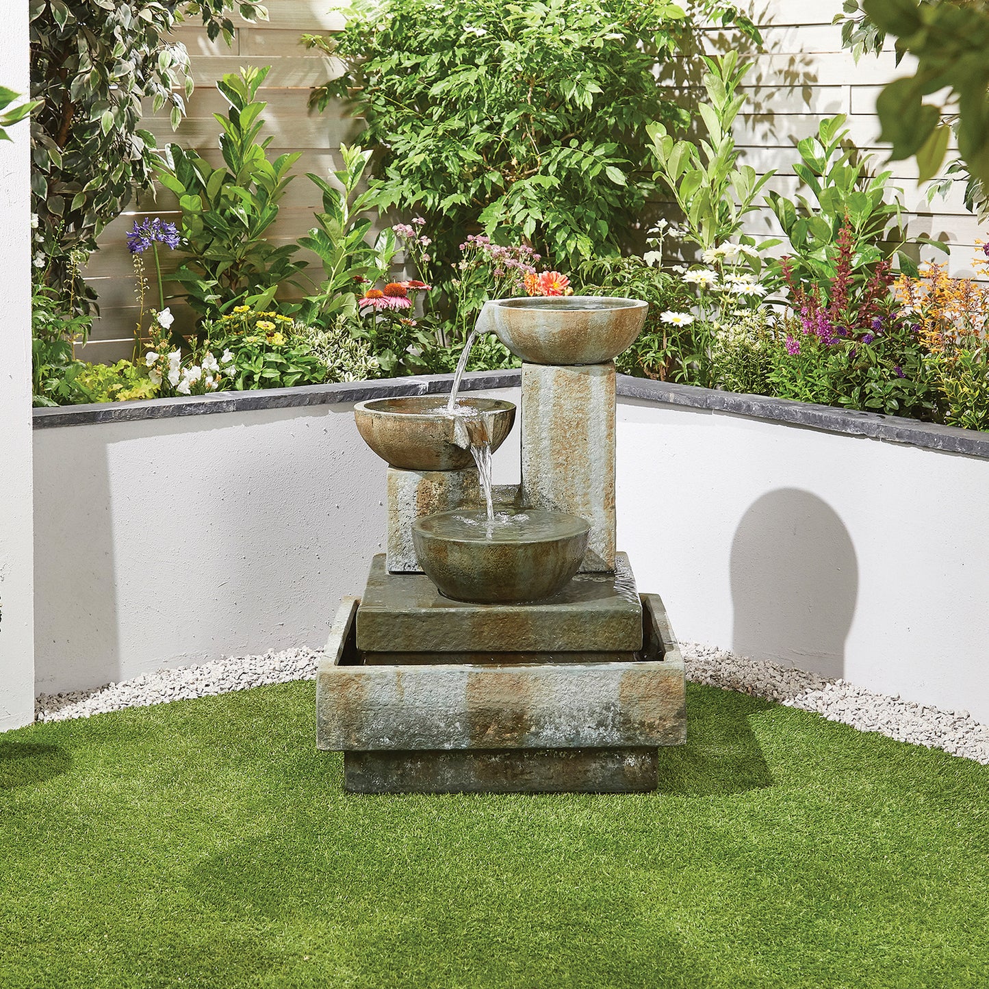 A tiered fountain designed by La Hacienda, featuring their Patina Bowls made from glass fibre reinforced concrete, gracefully flows with water in a garden corner. The fountain is surrounded by lush green shrubs and vibrant flowers, while manicured grass and a patch of white pebbles cover the ground.