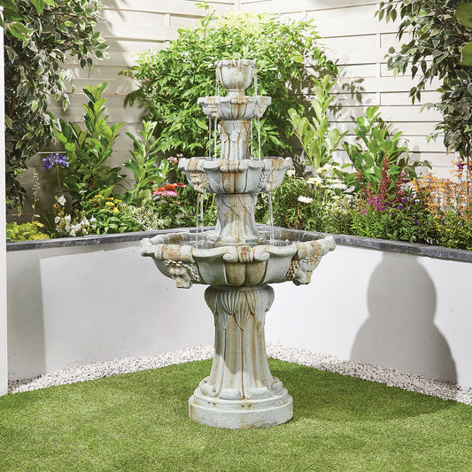 A three-tiered *La Hacienda* Lioness Fountain graces the garden, enveloped by lush greenery and vibrant flowers. Water gracefully cascades down each lion-inspired tier. Positioned against a white fence, this fountain is securely anchored with reinforced concrete and is surrounded by grass with a small gravel border at its base.