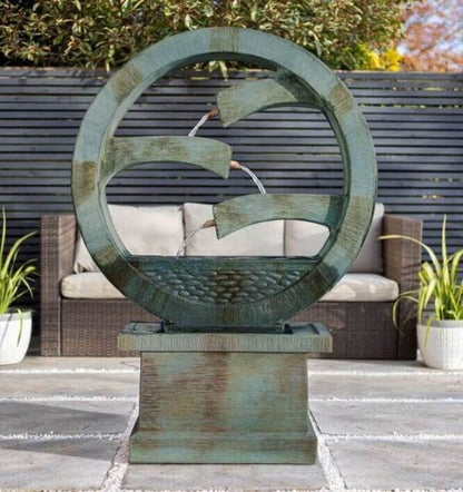 The La Hacienda Tranquil Spills is a circular, tiered contemporary fountain crafted from stone, featuring serene streams of water cascading through three curved channels. It elegantly rests on a square pedestal in front of a wicker sofa, with a backdrop of dark slatted fencing and potted plants, making it an ideal centerpiece for any garden setting.