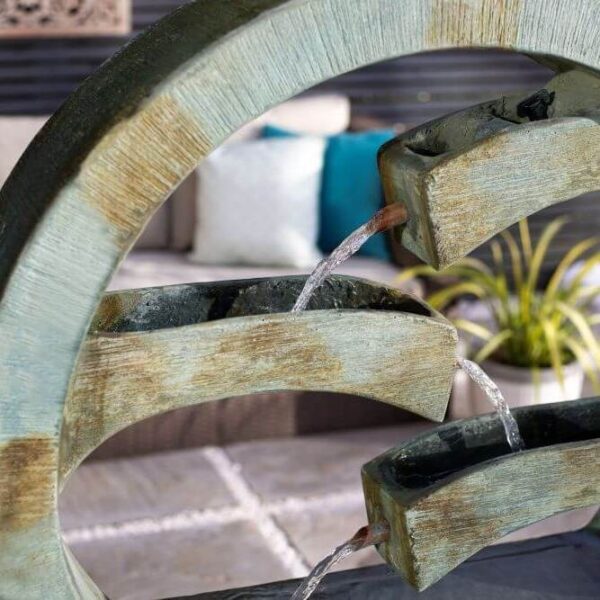 A close-up of the Tranquil Spills garden centerpiece, a contemporary fountain by La Hacienda featuring a circular design with a weathered stone finish. Water flows gently from each tier, creating soothing spills. In the blurred background, there is a seating area adorned with cushions and surrounded by plants.