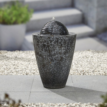 The La Hacienda Dappled Column, a modern dark stone fountain with a spherical top and a built-in recirculation pump, gently trickles water. It is bordered by white pebbles on a gray tiled surface. This contemporary setup includes steps and a potted plant in the blurred background.