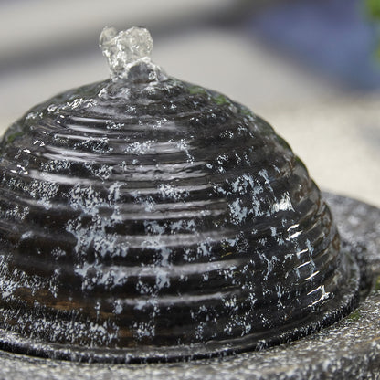 A close-up of the Dappled Column fountain by La Hacienda reveals its textured, dome-shaped design as water gracefully cascades over the ribbed surface. This modern piece features clear droplets shimmering on the dark material, against a blurred and abstract background.