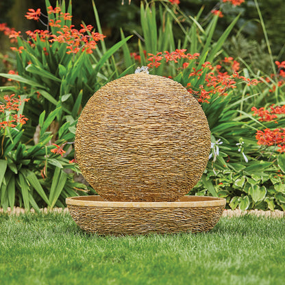 The Cotswold Sun, a spherical water feature by La Hacienda, is made from Glassfibre Reinforced Concrete. It is the centerpiece of a garden, where its textured stone surface is beautifully highlighted by the lush green grass and vibrant red flowers, representing a modern design.