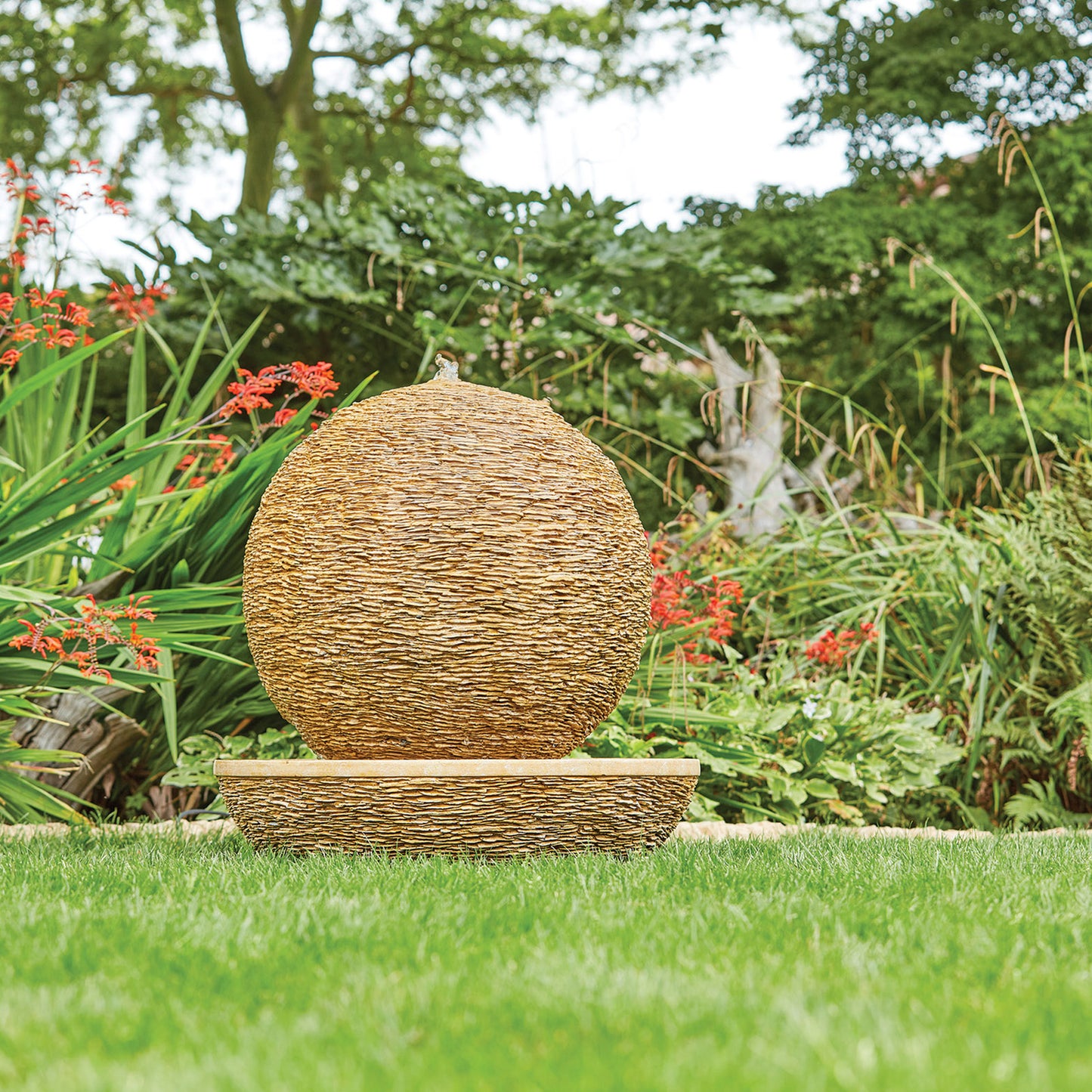 The Cotswold Sun by La Hacienda, a woven sphere sculpture made from Glassfibre Reinforced Concrete, rests on a flat base amidst a lush garden. It is surrounded by vibrant red flowers and diverse green plants, creating a serene outdoor setting with a modern design flair.