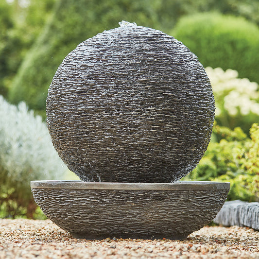 The Mysterious Moon by La Hacienda, a large spherical stone water feature made from glassfibre reinforced concrete, is positioned on a matching basin surrounded by gravel and lush greenery. This self-contained piece allows water to cascade down its textured surface, enhancing the soothing outdoor aesthetic.