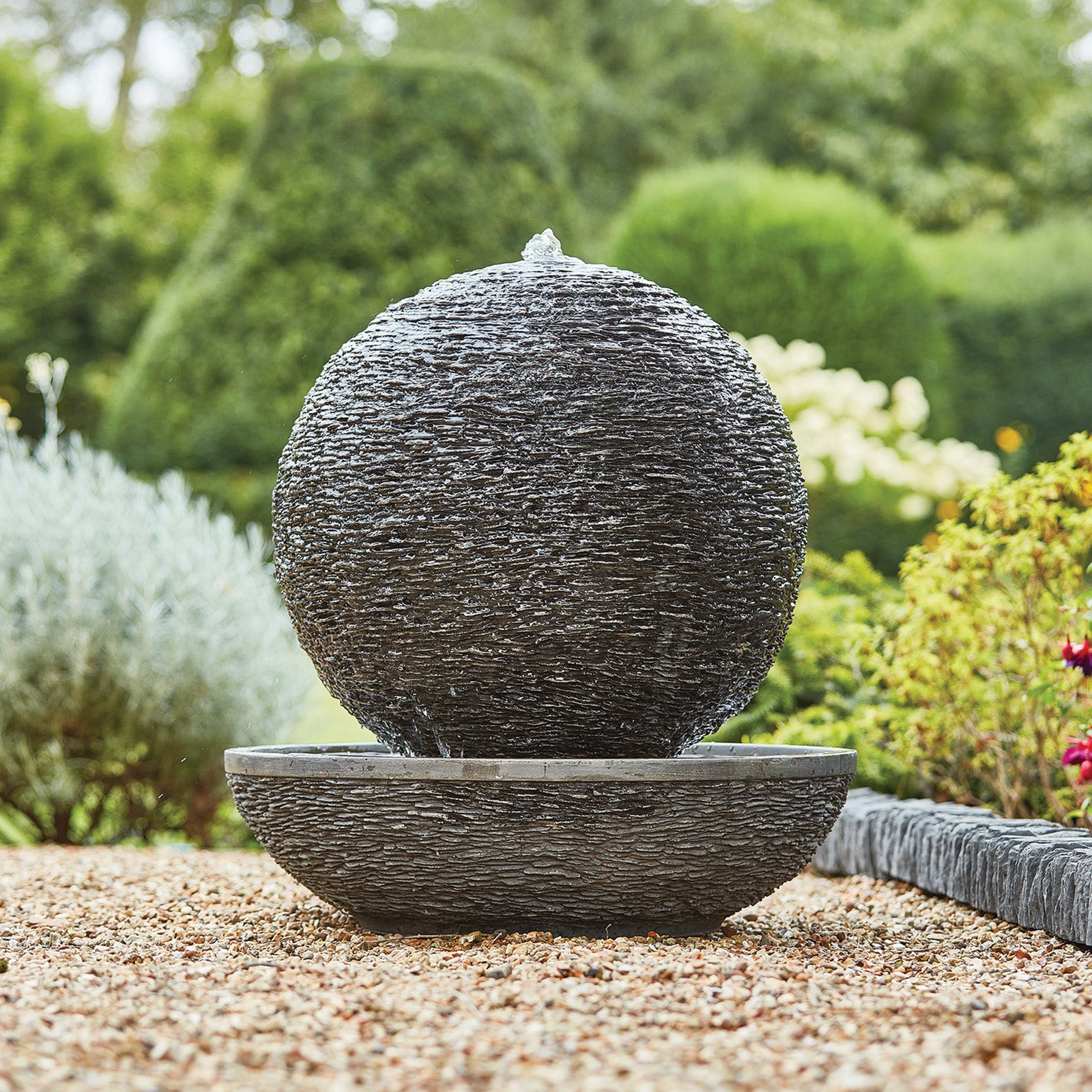 The Mysterious Moon by La Hacienda, a round stone water feature made from Glassfibre Reinforced Concrete, is placed on a gravel surface in the garden. Water flows gently over its textured exterior into a shallow basin below, while the background of lush greenery and manicured shrubs completes this tranquil self-contained oasis.