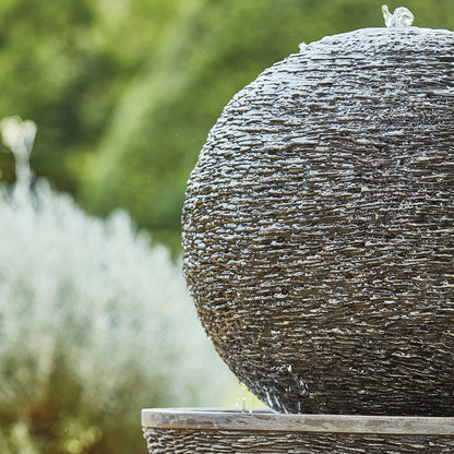 The image showcases a close-up of the La Hacienda Mysterious Moon, a spherical stone fountain made from glassfibre reinforced concrete, with water gracefully cascading over its textured surface. The softly blurred background highlights lush green foliage, enhancing the serene ambiance of the garden setting.